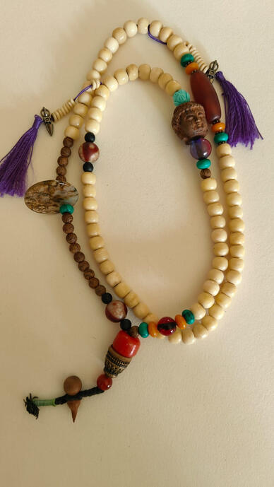 Embrace the spirit of wanderlust with this one-of-a-kind handmade mala necklace. Featuring a serene wooden Buddha head, creamy natural beads, vibrant gemstone accents, and enchanting purple tassels, this piece is perfect for layering or wearing as a bold s